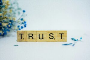 The word "TRUST" is spelled out with Scrabble letters in front of a background with flowers.