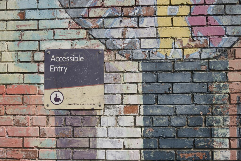 Accessible entry sign with a wheelchair symbol mounted on a colorful, weathered brick wall.