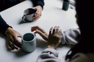 Two people in a comfortable relationship seated at a table with coffee cups, gesturing as they talk during a conversation.