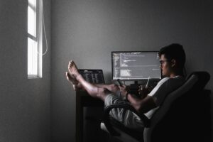 A gig developer sits with feet up, phone in hand, casually working on a home computer setup.