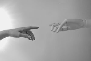 A human hand and a robotic hand reach toward each other against a grey background, evoking the tension and relationship between human creativity and artificial intelligence in modern marketing.