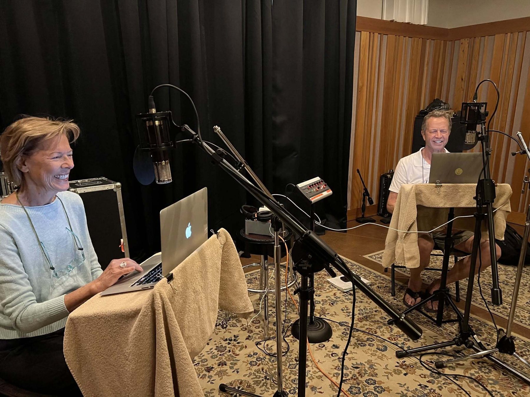 Two narrators reading from laptops at studio microphones during an audiobook recording session