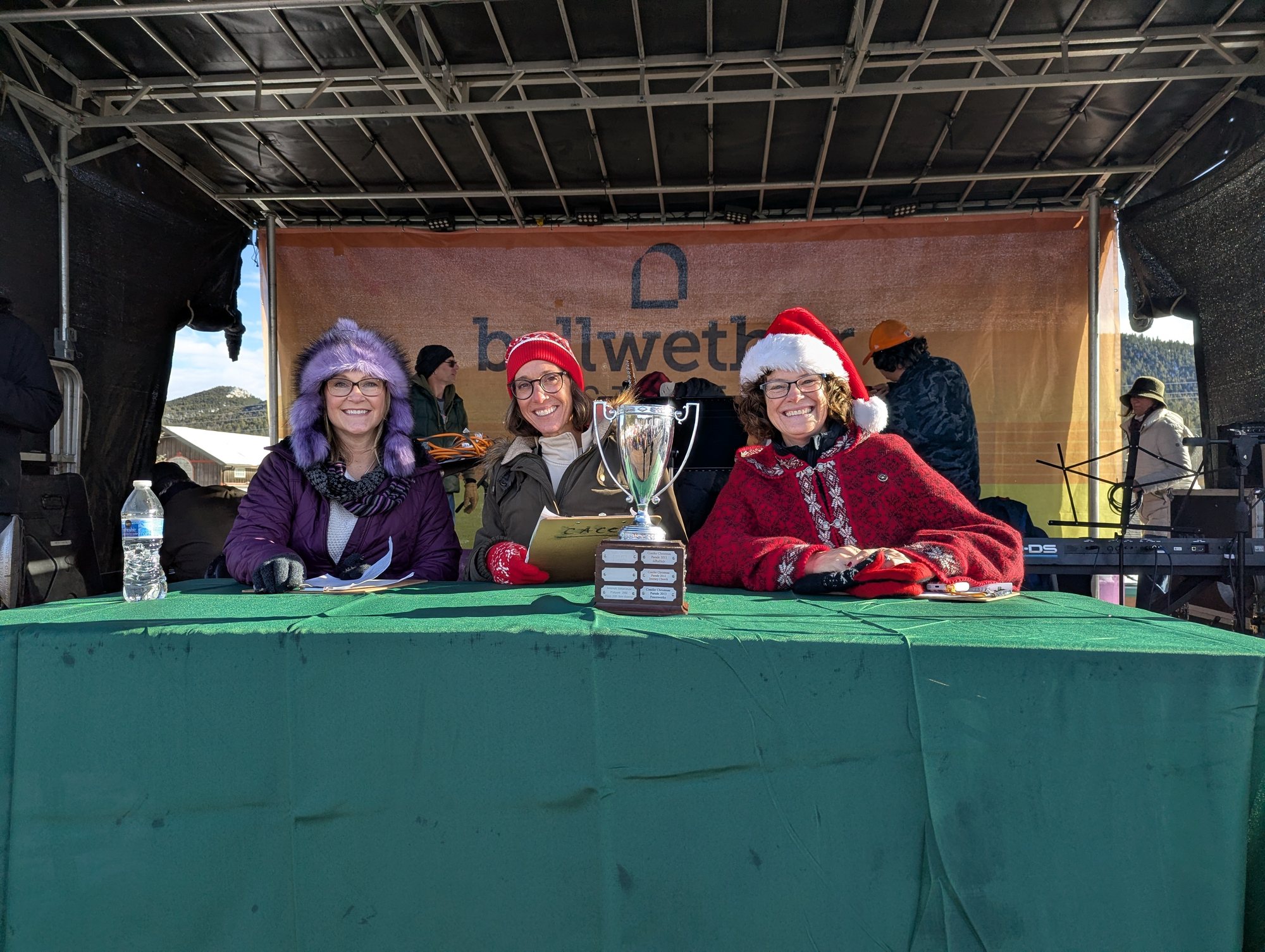 Three parade judges in winter and Santa hats seated at a green table on an outdoor stage, with a trophy in front of them
