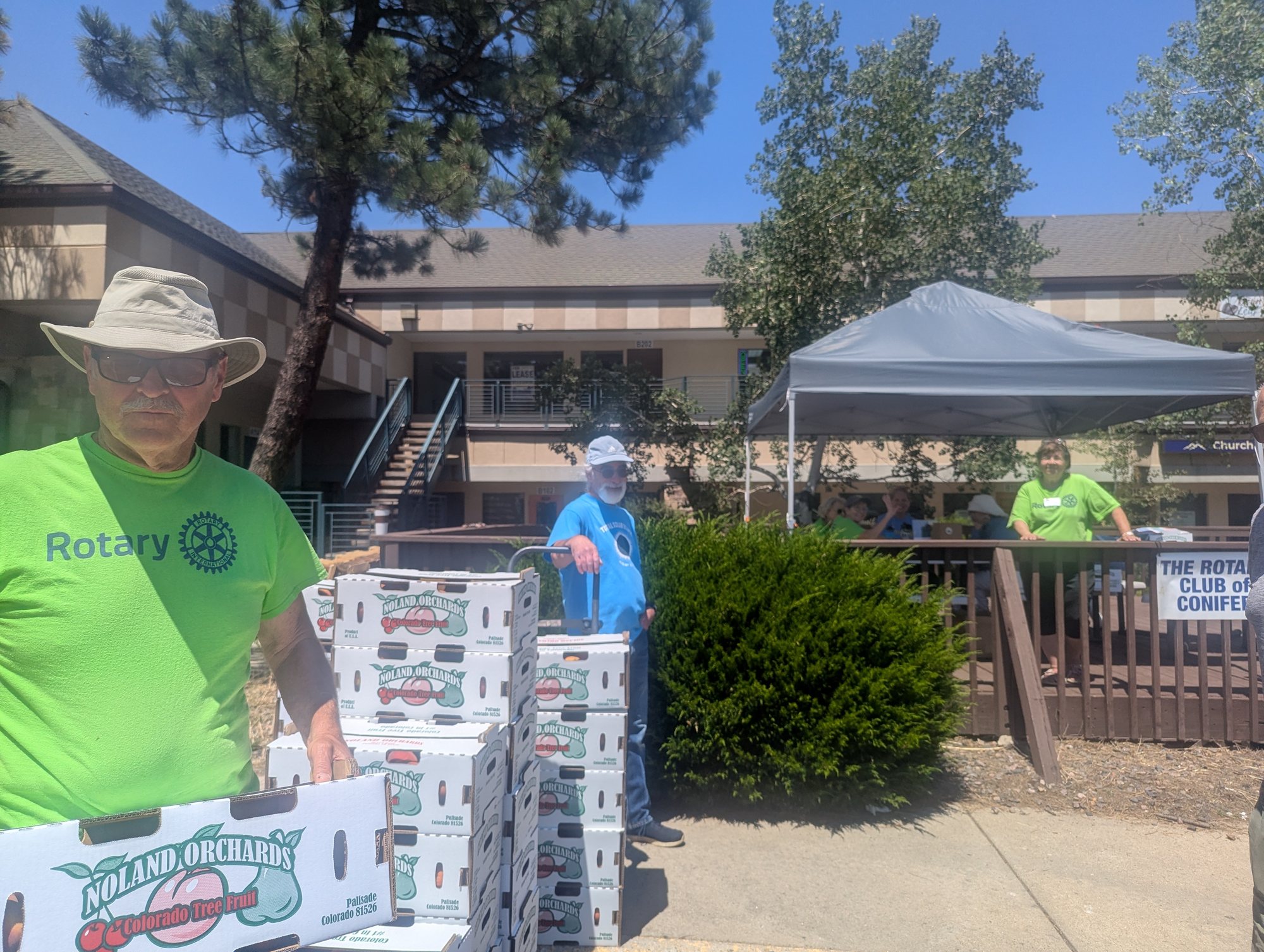 A Rotary volunteer in a green shirt beside stacked boxes of Noland Orchards peaches outside a shopping center