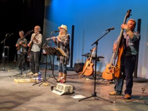 A folk band with guitars, a flute and an upright bass playing on a stage lit in blue