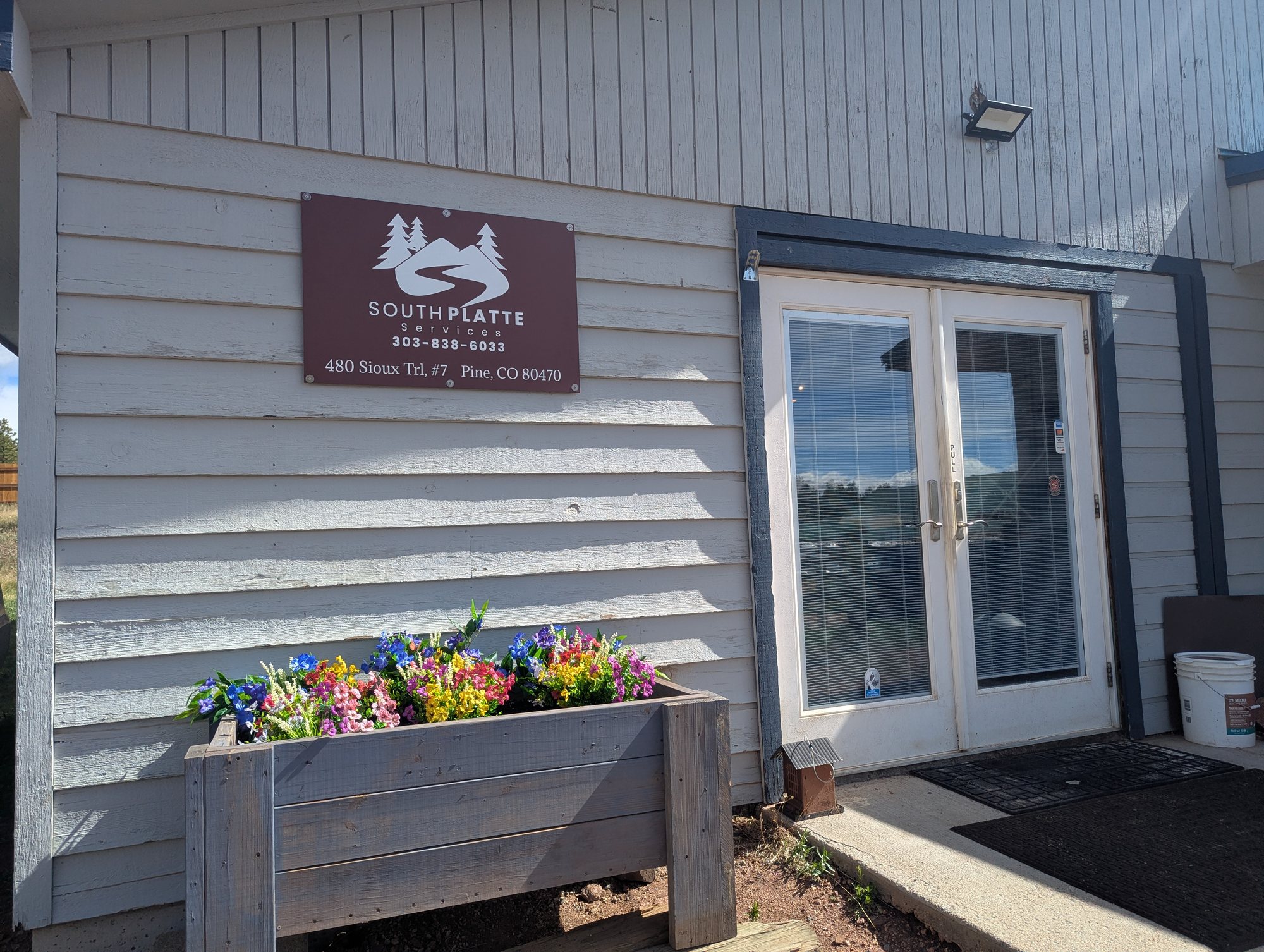 A gray office building with a maroon South Platte Services sign above a planter box of bright flowers beside glass double doors
