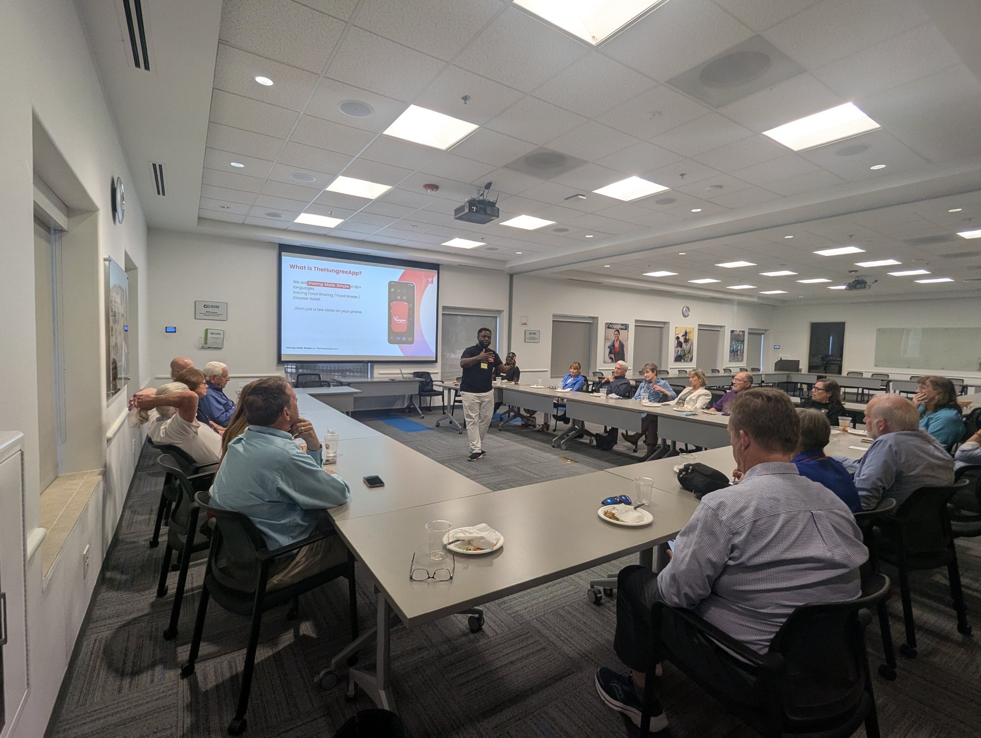 A speaker presenting to a seated group around long tables in a bright conference room