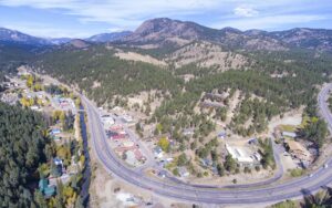An aerial view of a small mountain town strung along a curving highway and a river, with pine-covered hills and peaks behind