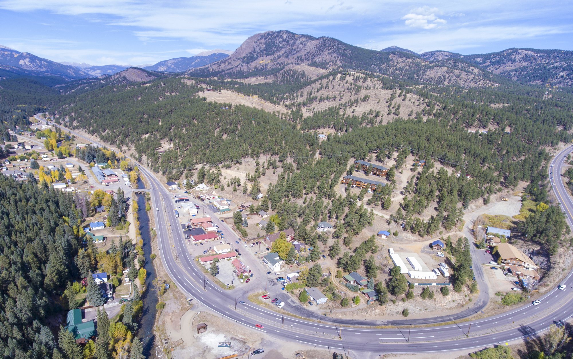 An aerial view of a small mountain town strung along a curving highway and a river, with pine-covered hills and peaks behind