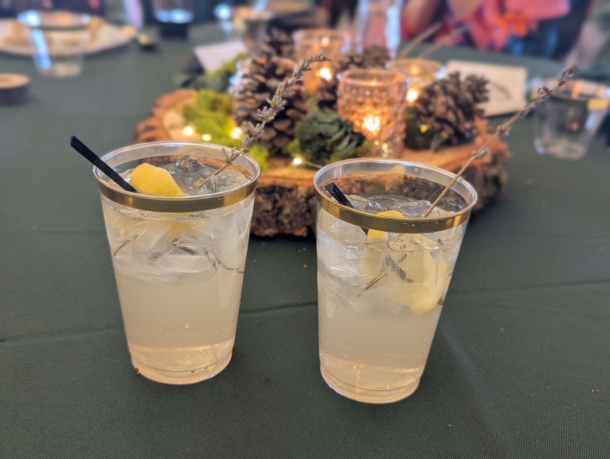 Two glasses of lemonade garnished with lemon and lavender sprigs on a green tablecloth, with a candlelit pinecone centerpiece behind