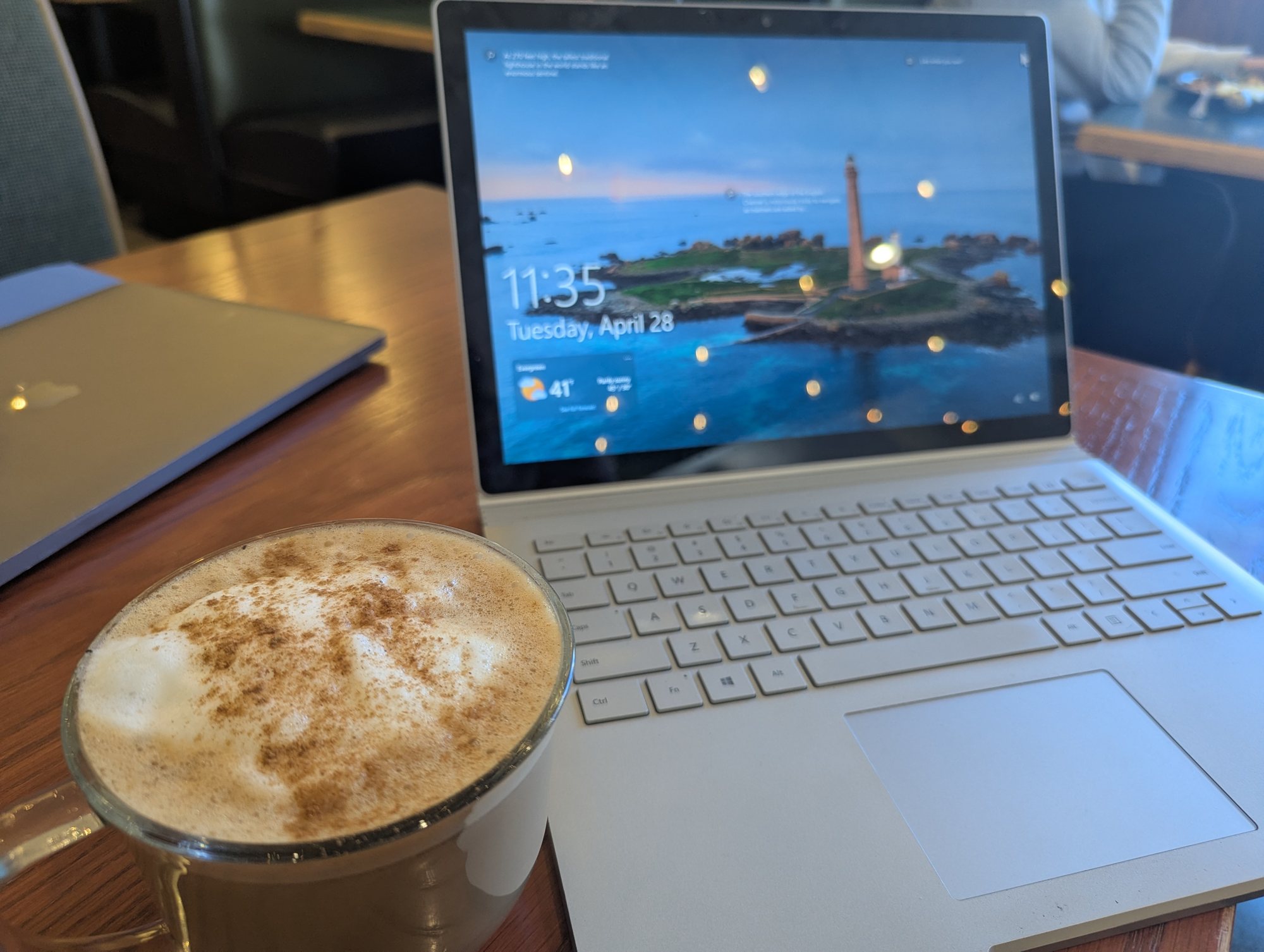 A cinnamon-dusted cappuccino in a glass mug in front of an open laptop on a wooden cafe table
