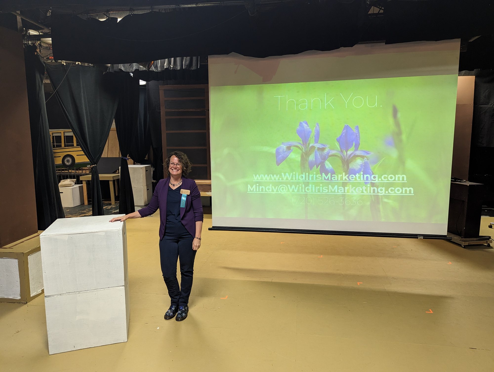 A smiling woman in a purple jacket on a stage beside a projected thank-you slide with blue iris flowers and the Wild Iris Marketing web address