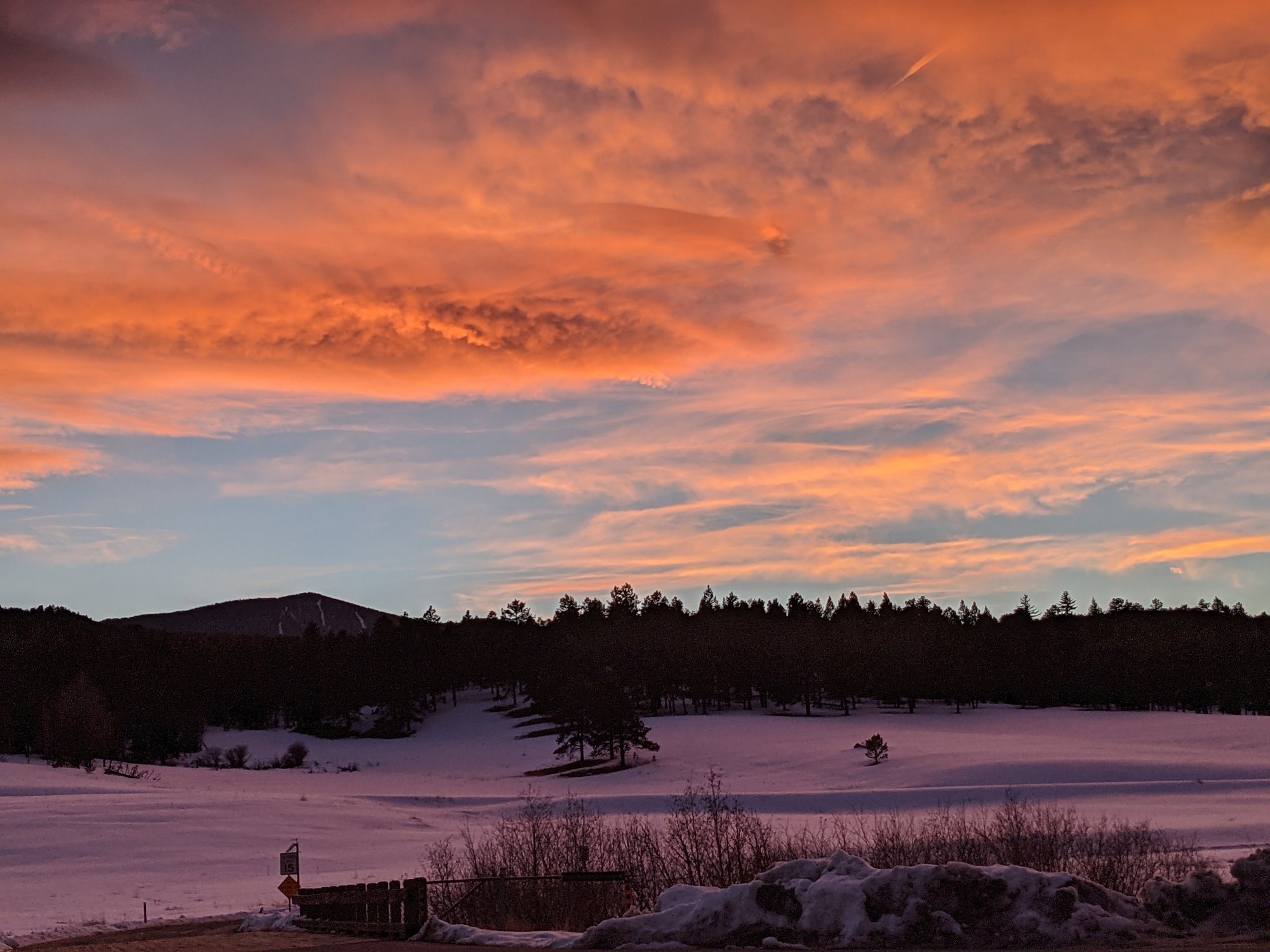 Orange and pink clouds over a snow-covered meadow ringed by dark pines