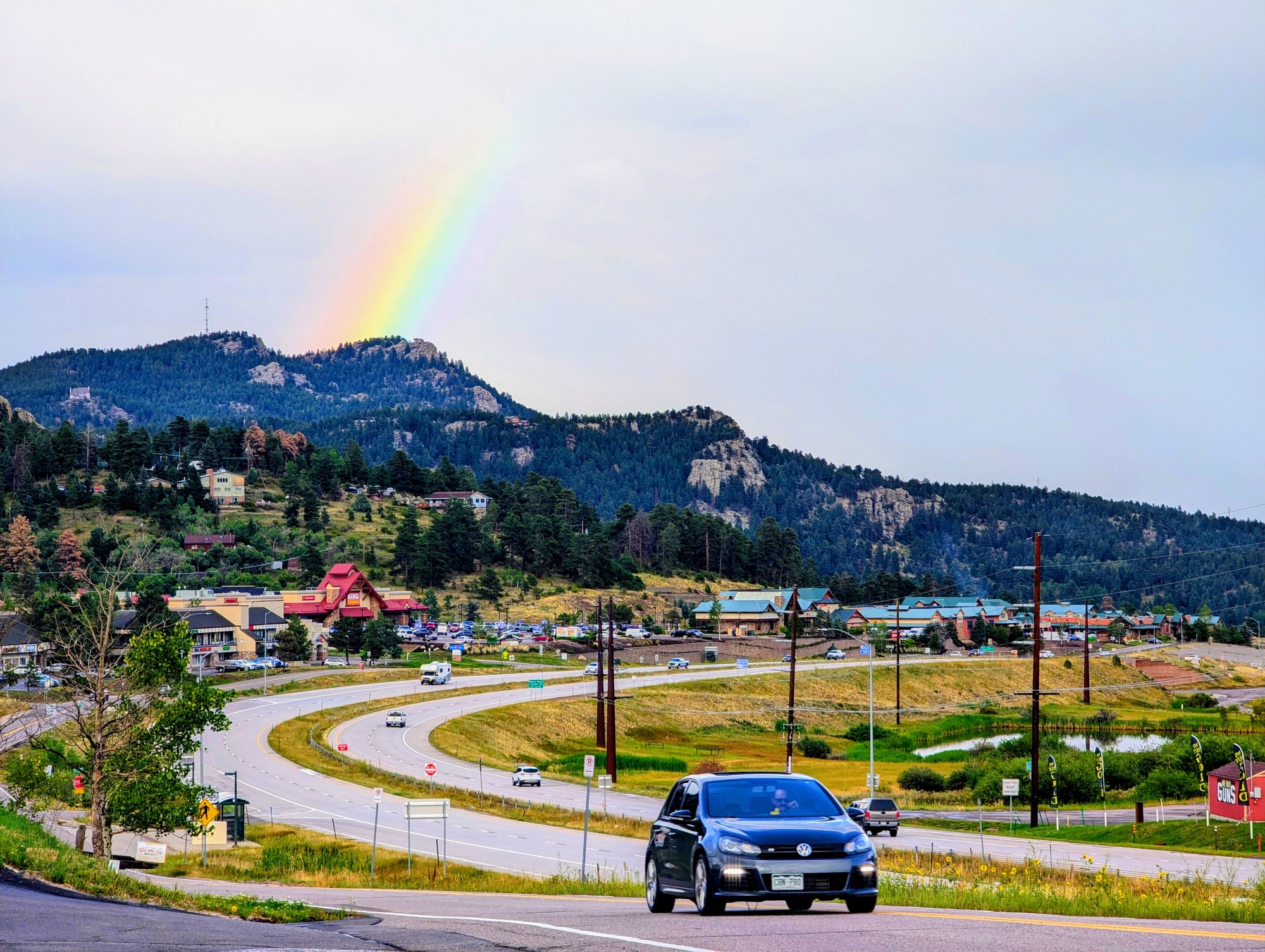 A rainbow over a rocky, pine-covered hill above a curving highway and the shops of Conifer