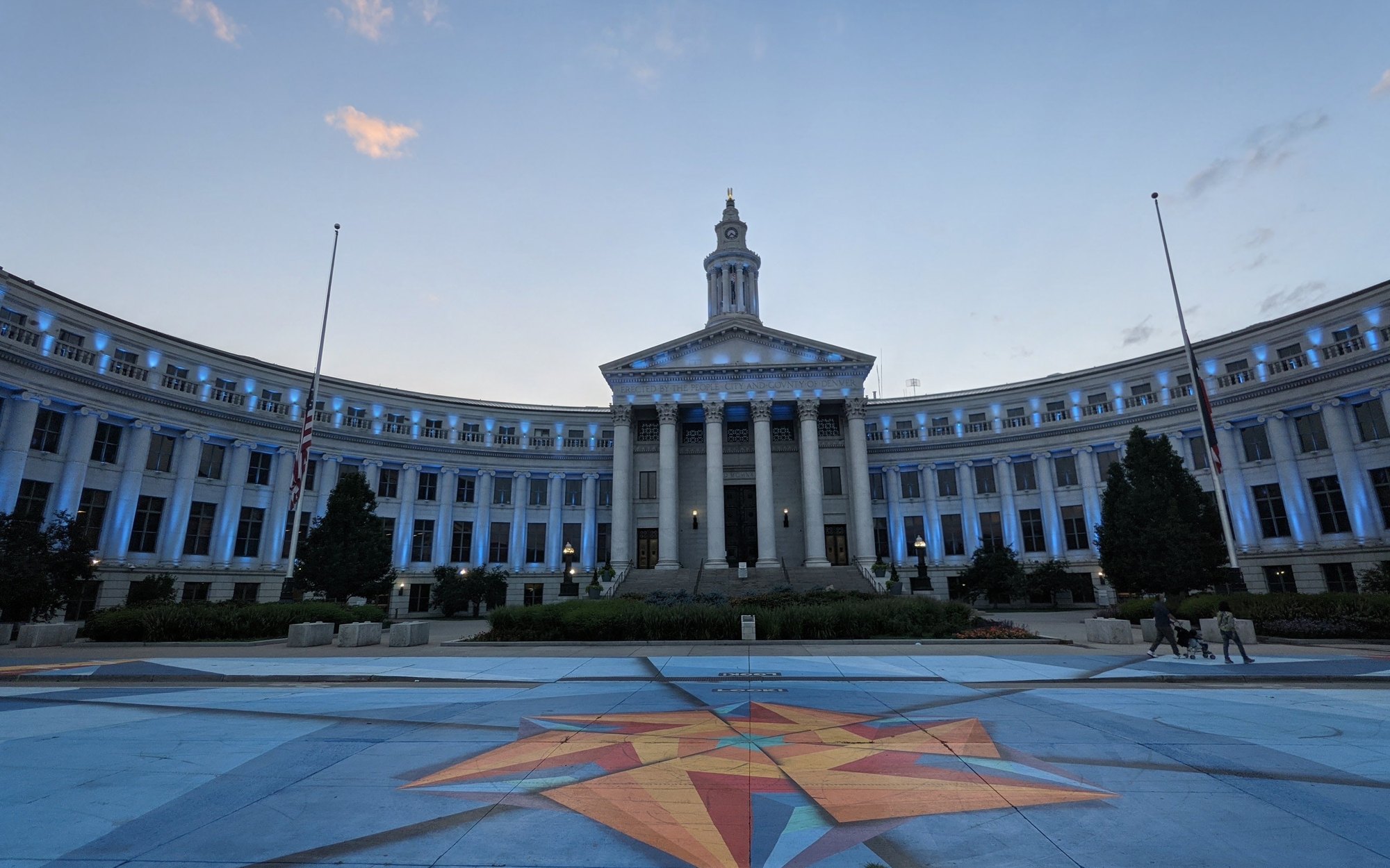 The Denver City and County Building at dusk, its curved colonnade and clock tower lit in teal, with a painted compass star on the plaza in front.