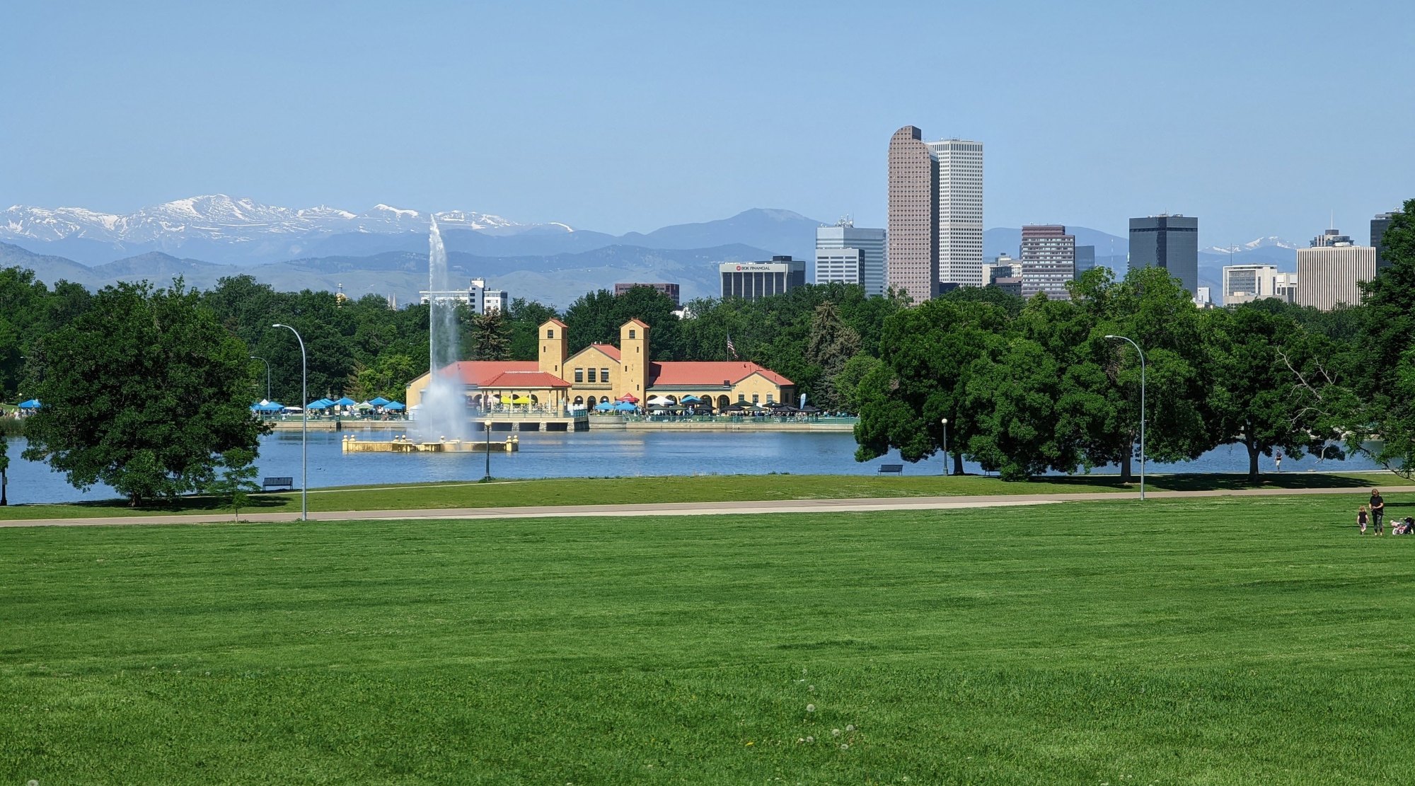 Denver's downtown skyline and snow-capped mountains seen across City Park, with a fountain in Ferril Lake and the yellow, red-roofed City Park Pavilion by the water.
