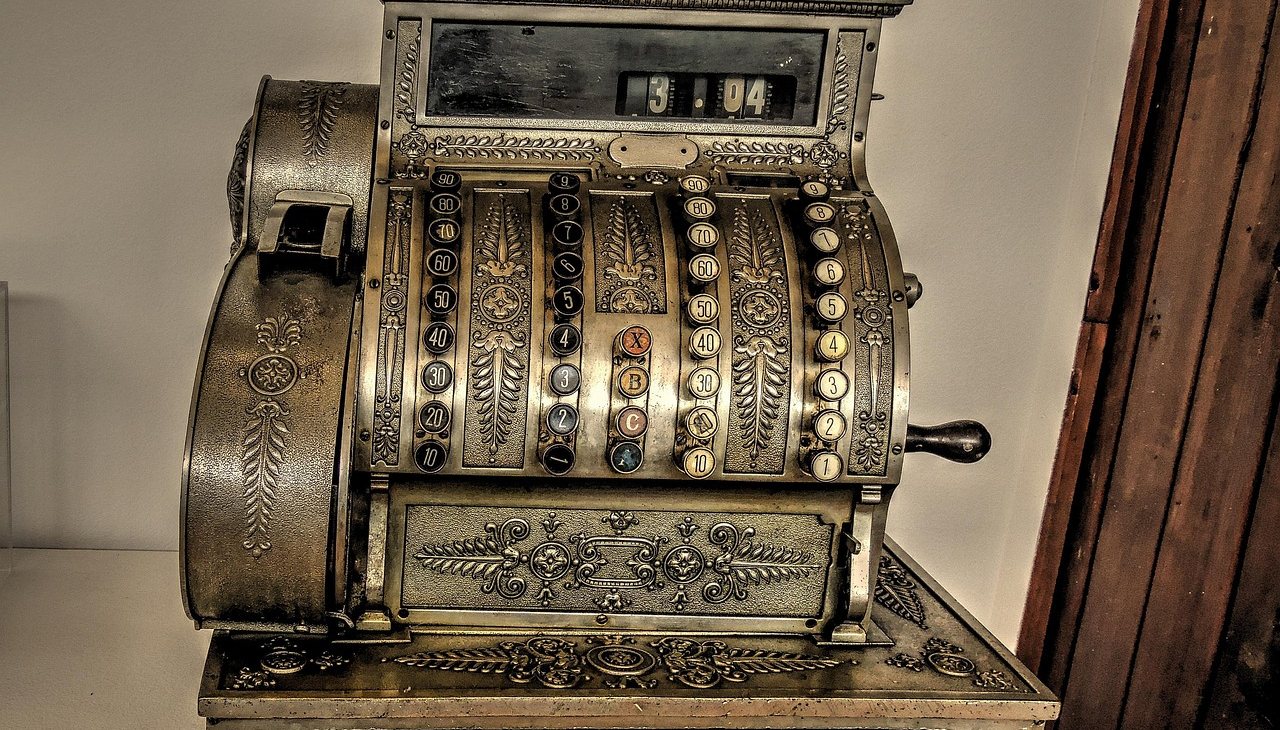 An ornate antique brass cash register with rows of round number keys, a side crank and a decorated drawer front