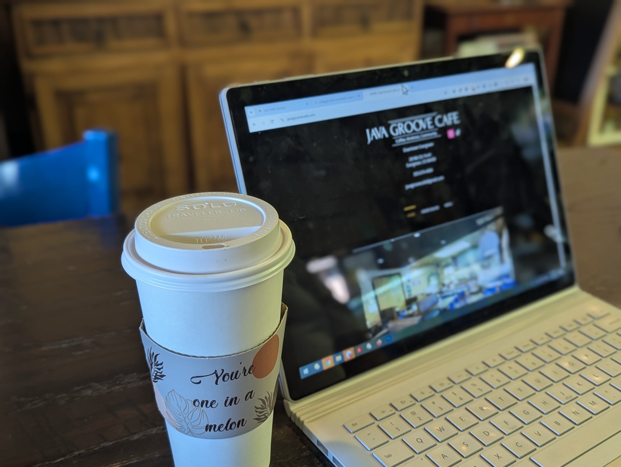 A to-go coffee cup in a sleeve reading "You're one in a melon" beside an open laptop on a cafe table