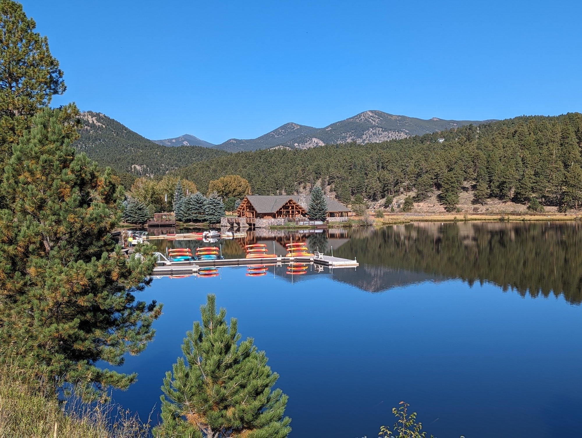 A log lake house and colorful boats on Evergreen Lake, reflecting pine-covered hills under a blue sky