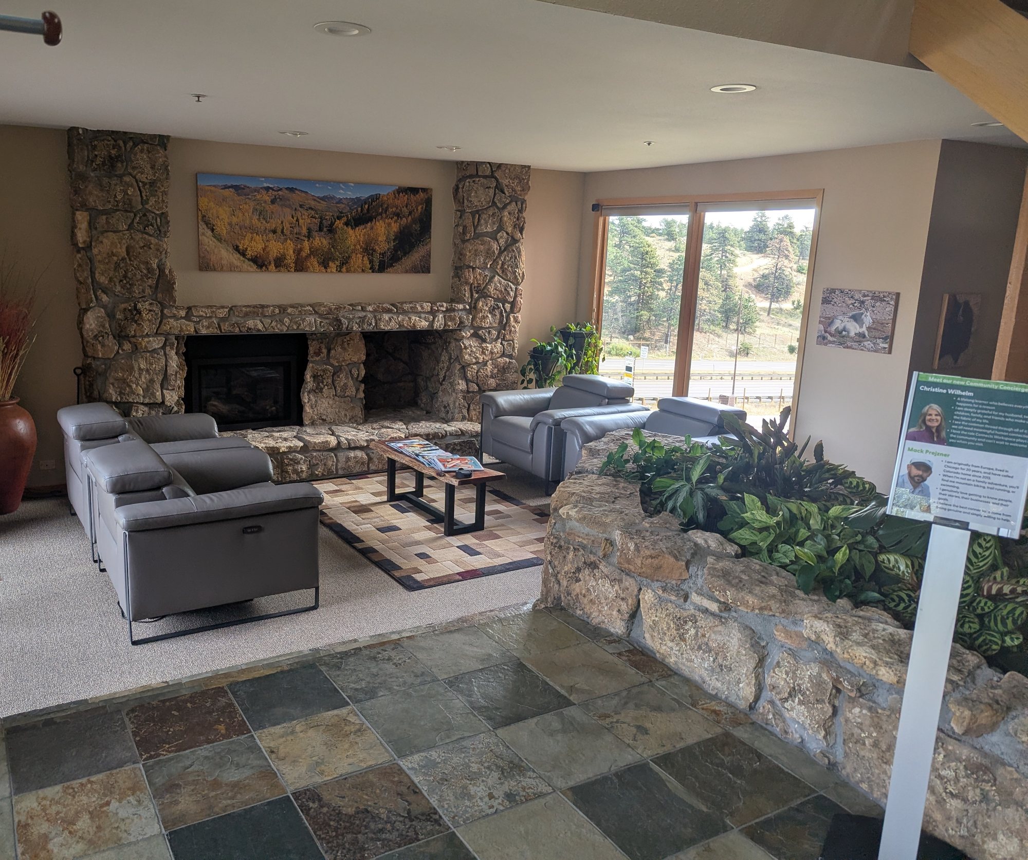 A bright lobby with gray leather chairs, a stone fireplace, a large aspen painting and a window onto pine trees