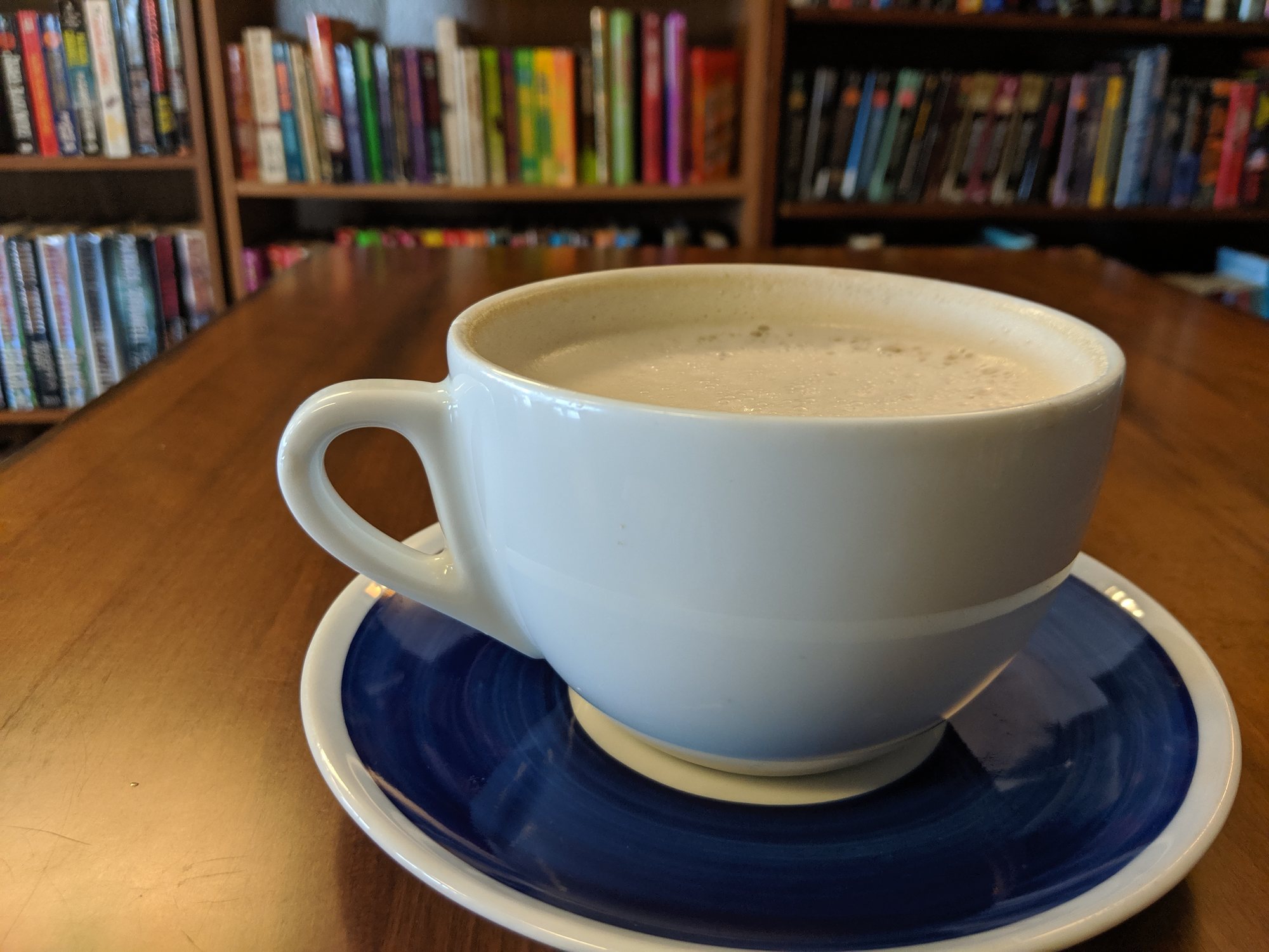 A white cup of latte on a blue saucer on a wooden table, with shelves of books behind it