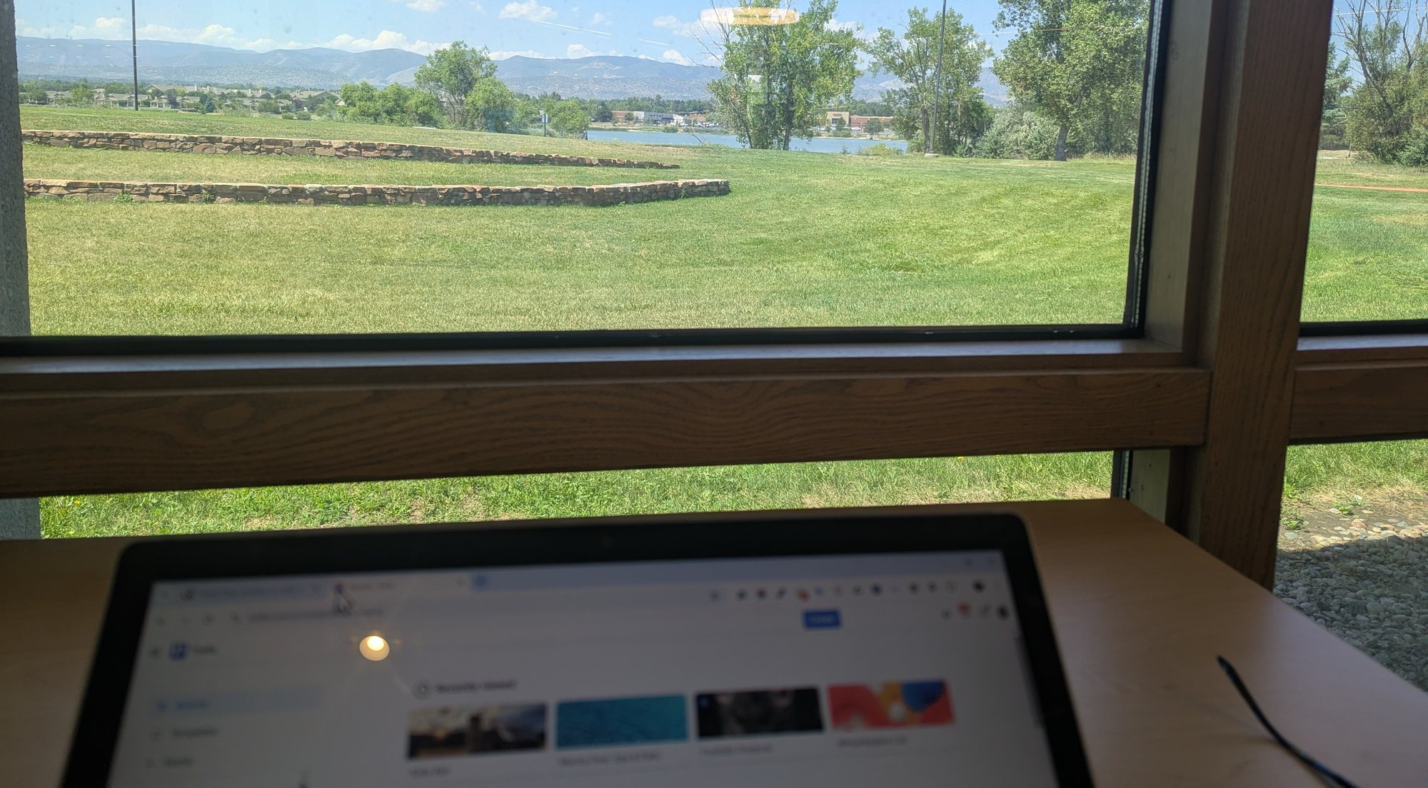 A laptop on a table by a big window looking out at a green lawn, trees and the foothills