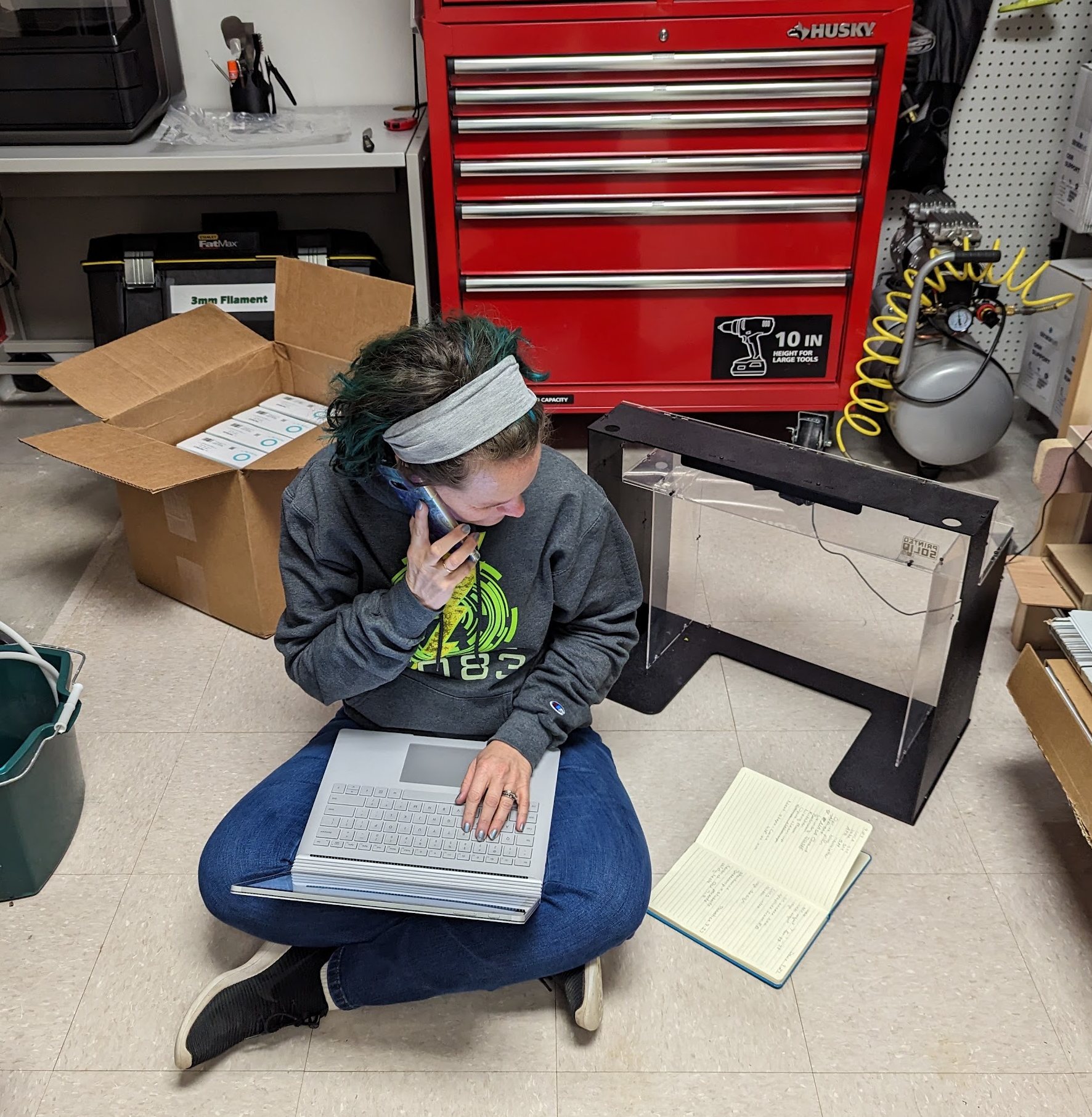 A woman sitting cross-legged on a workroom floor with a laptop on her lap and a phone to her ear, surrounded by tool chests, boxes and robot parts