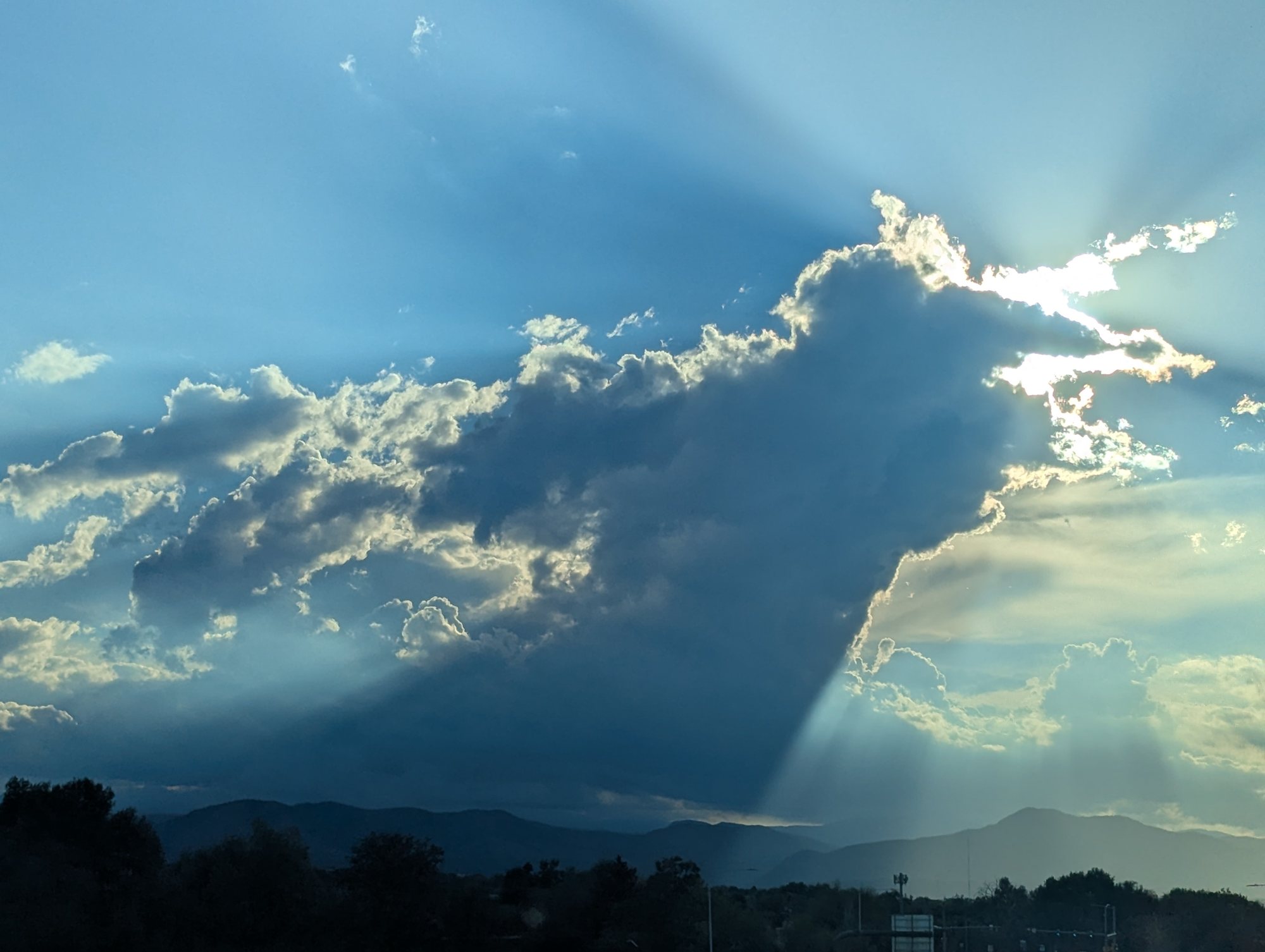 Rays of sunlight streaming from behind a large cloud over a dark line of foothills