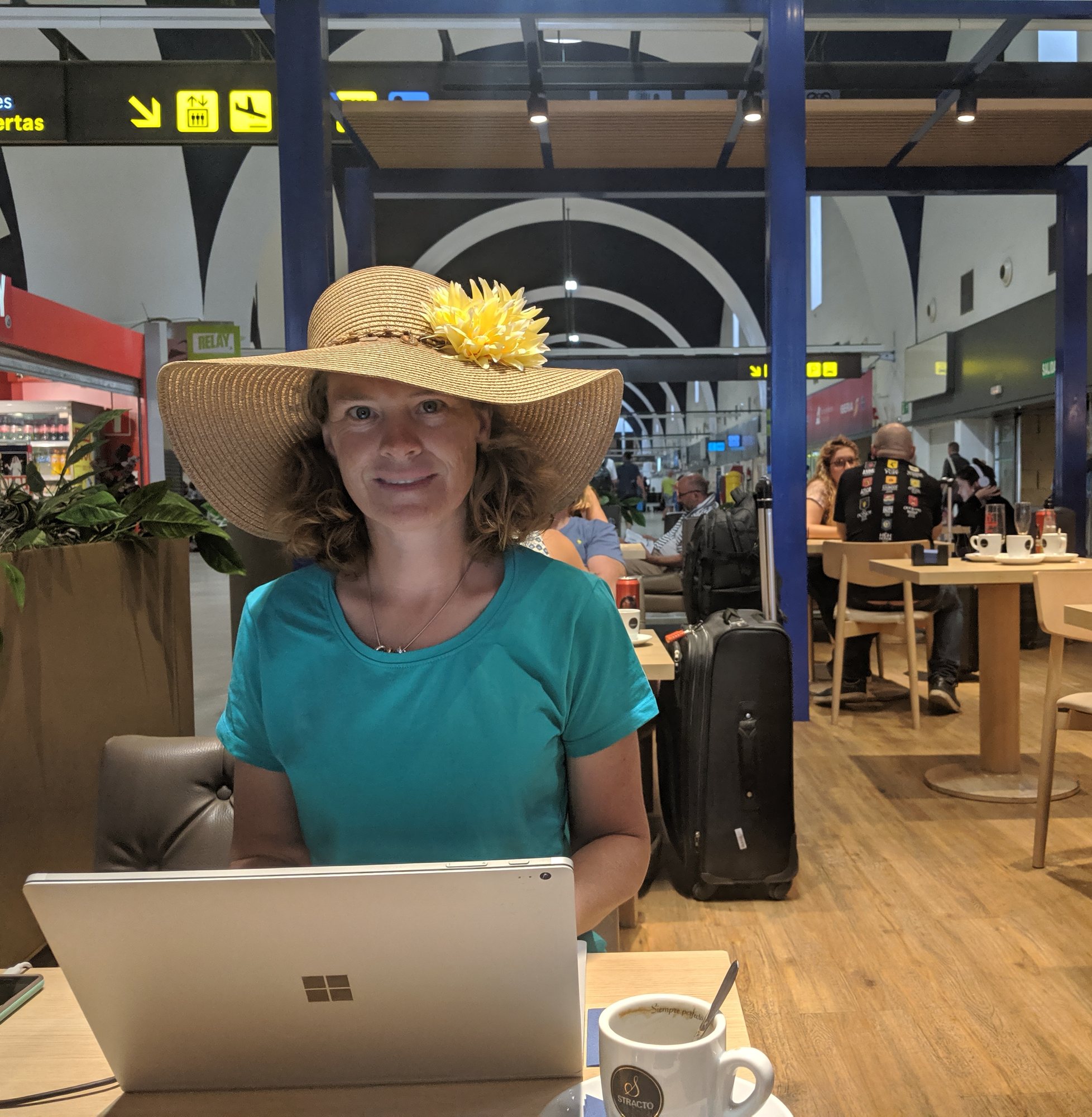 A woman in a wide sun hat and teal shirt working on a laptop at an airport cafe table, with a coffee cup in front of her