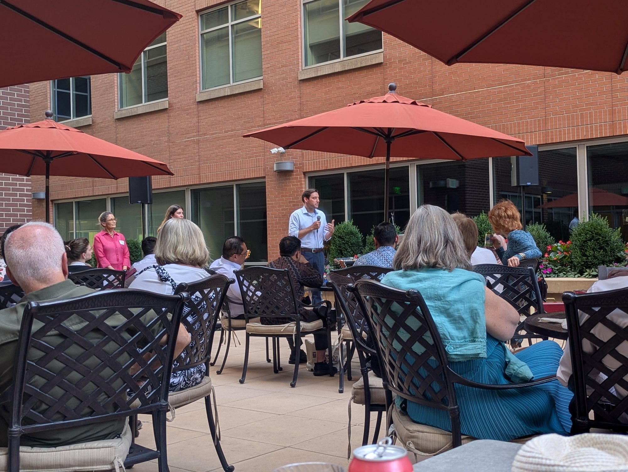 A speaker addressing guests seated under red patio umbrellas in a brick courtyard