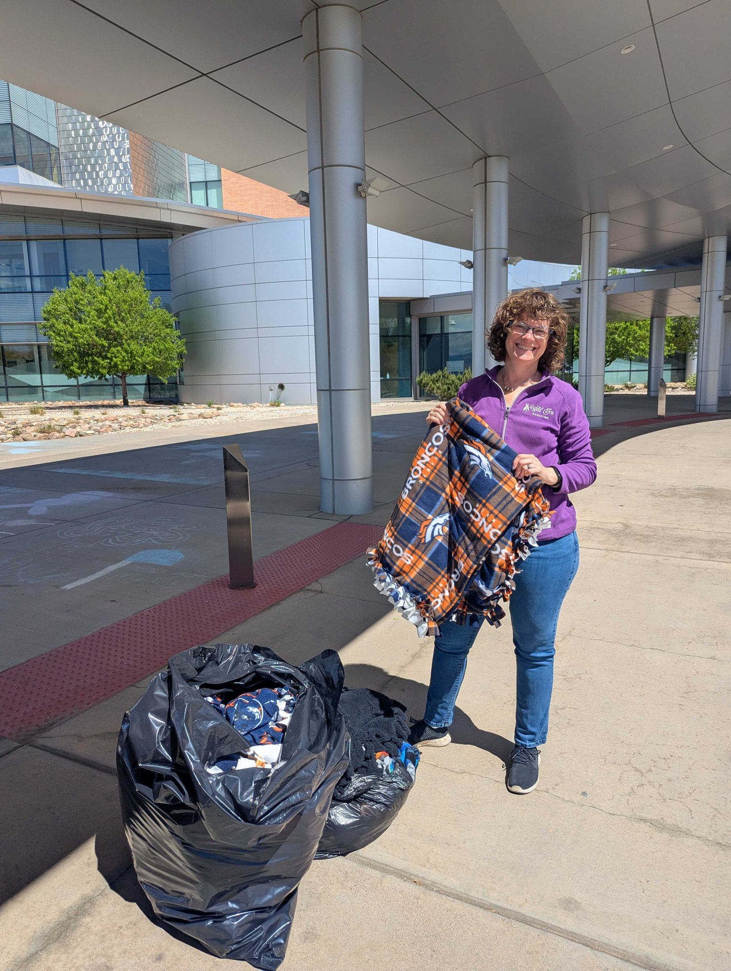 A smiling woman in a purple jacket holding up a Broncos fleece blanket beside bags of blankets outside a hospital entrance