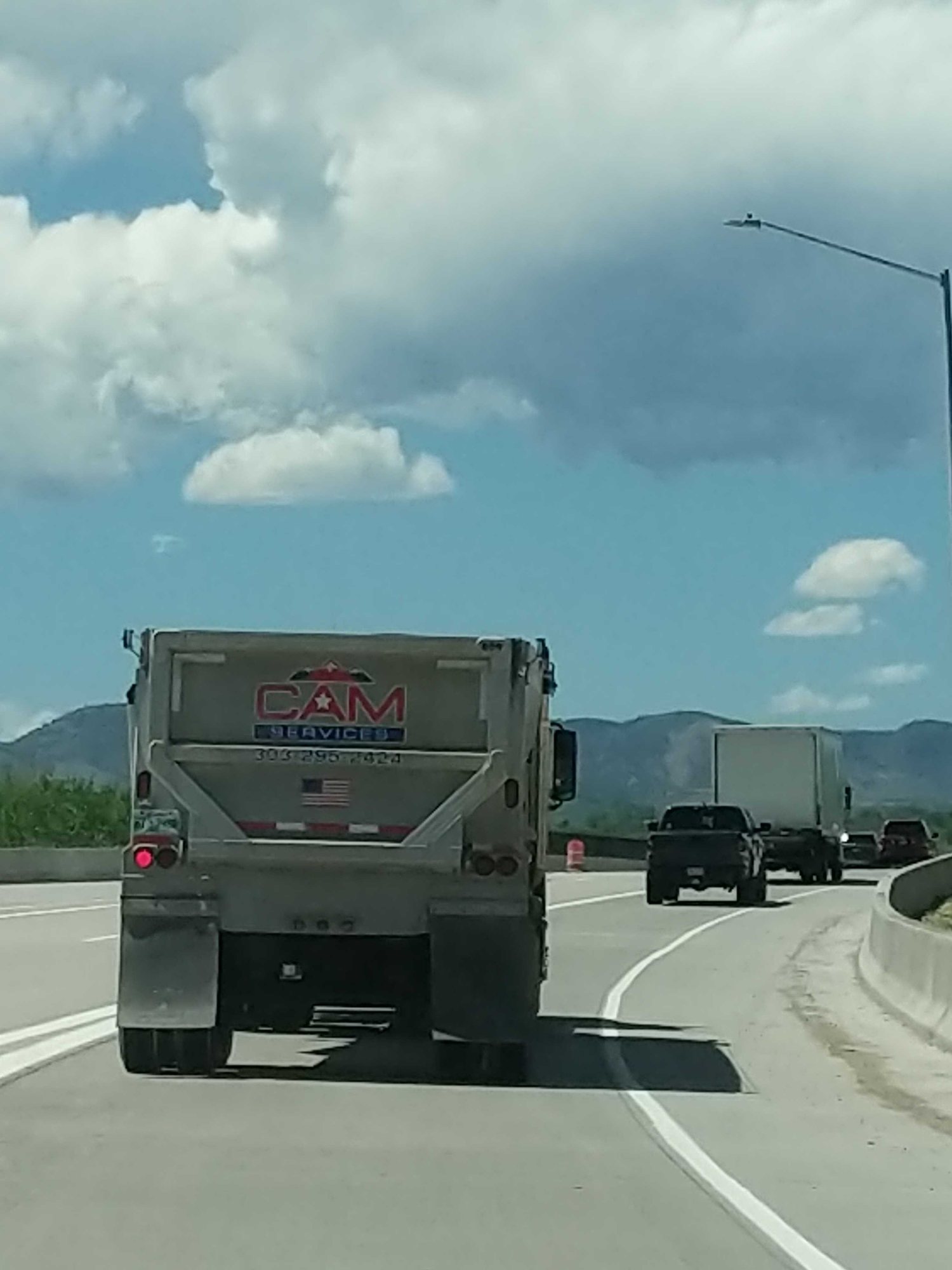 A dump truck with a company logo driving on a highway, with foothills and big clouds ahead