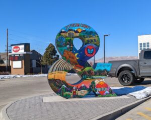 A large painted sculpture shaped like an ampersand, covered in murals of Jefferson County and Littleton scenes, under a blue sky